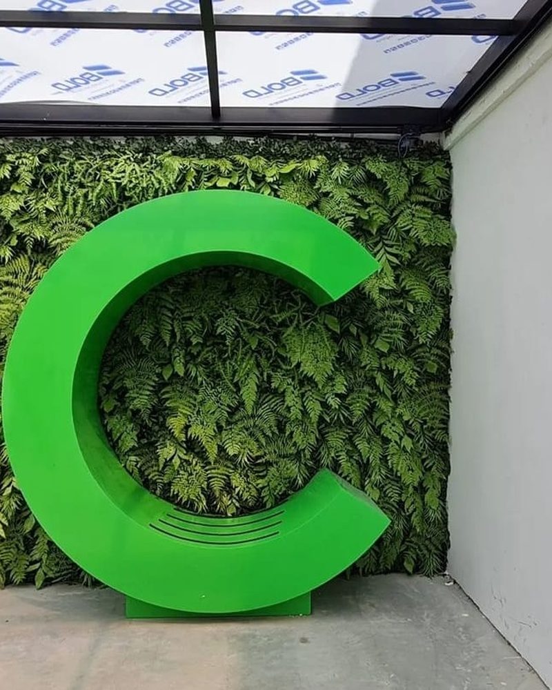 Peça cenográfica com logo 3D sobre jardim vertical de plantas artificiais