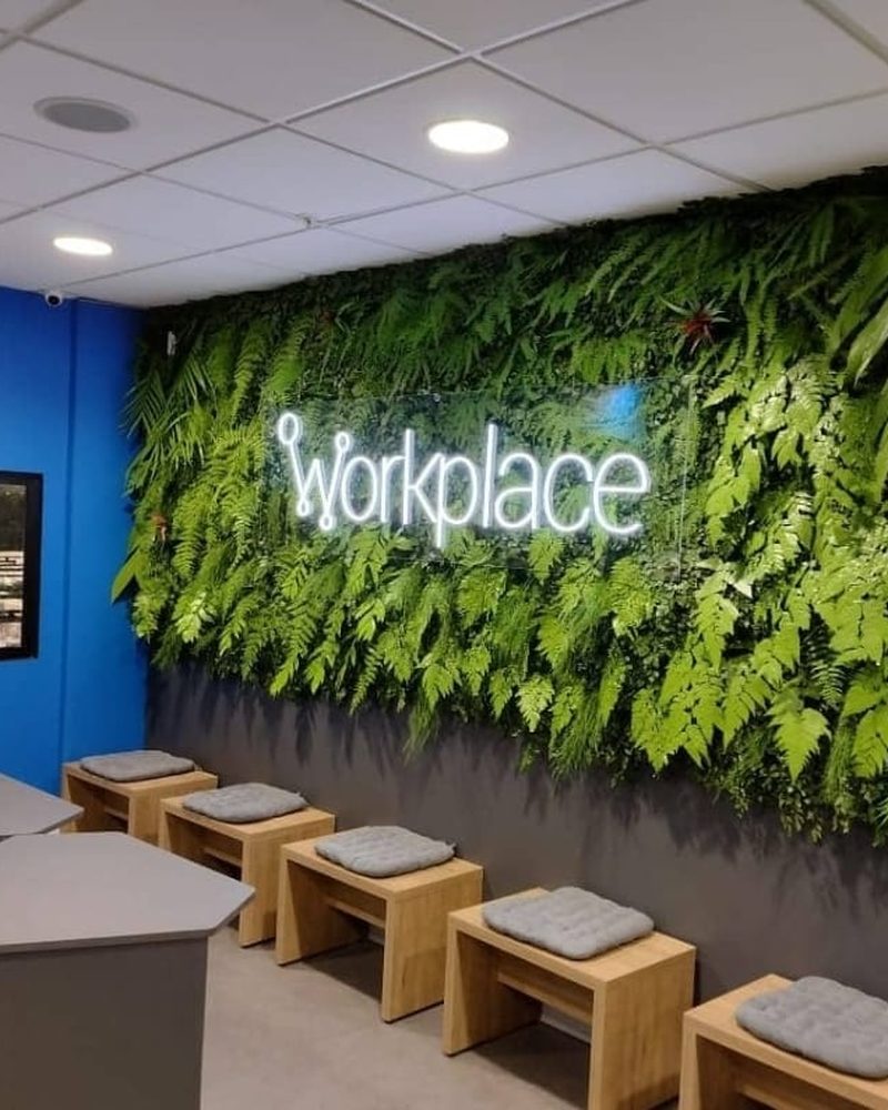 Sala de descanso com painel de plantas artificiais e letreiro neon workplace